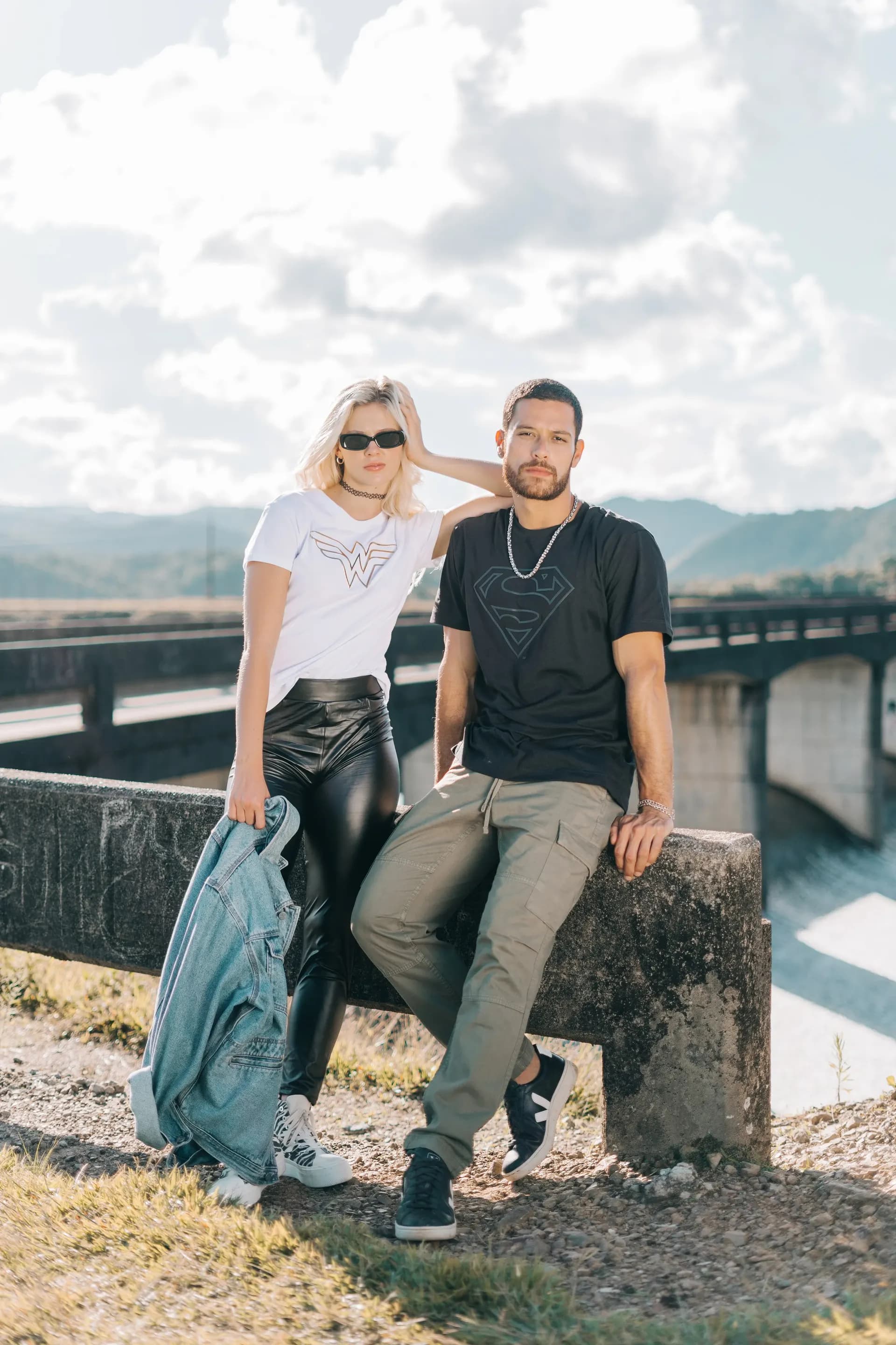 Retrato de la campaña TU PAR: pareja apoyada en un muro con un puente al fondo, con camisetas de Wonder Woman y Superman.
