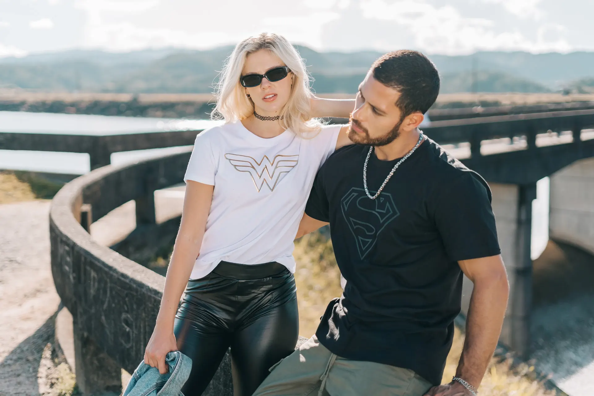 Fotografía editorial de la campaña TU PAR: pareja sobre un puente curvo de concreto, ella con camiseta de Wonder Woman y gafas de sol.