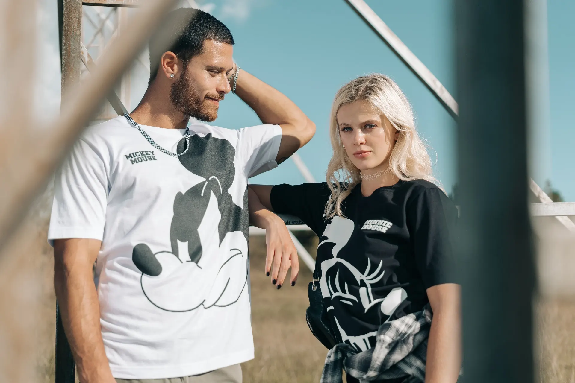 Fotografía editorial de la campaña TU PAR: pareja junto a una estructura metálica, él con camiseta blanca de Mickey Mouse y ella con camiseta negra.